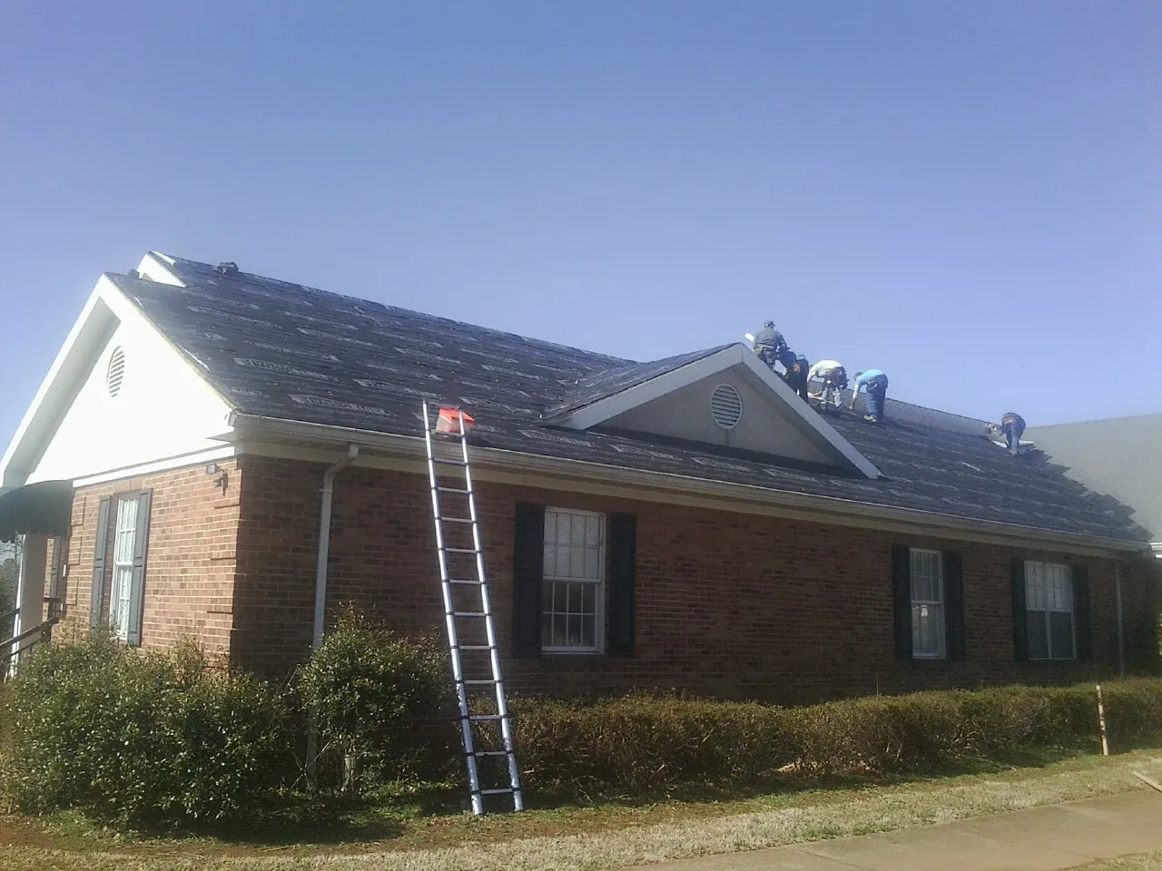 Crew tearing shingles off a brick ranch roof with a ladder set — Metal Roof Repair work by our crew in Pine Mountain