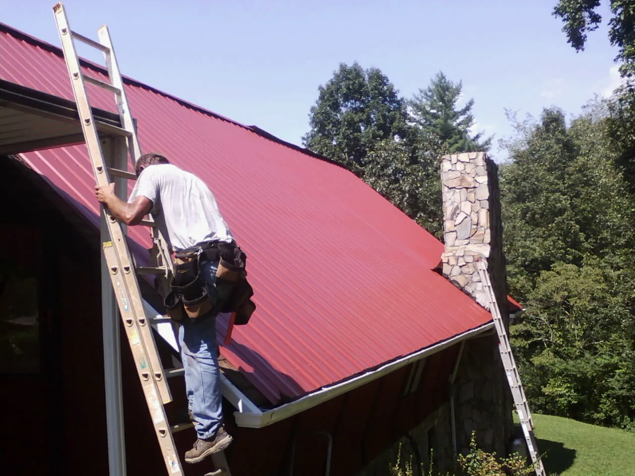 Roofer on a ladder beside a red metal roof and stone chimney — Soffit & Fascia Repair by Roofing Brothers in Pine Mountain